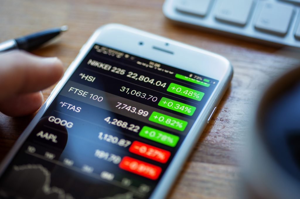 An Apple iPhone 6 on a desk displaying stock market information using the Apple Stock app. A human hand hovers above the FTSE 100 index figure.