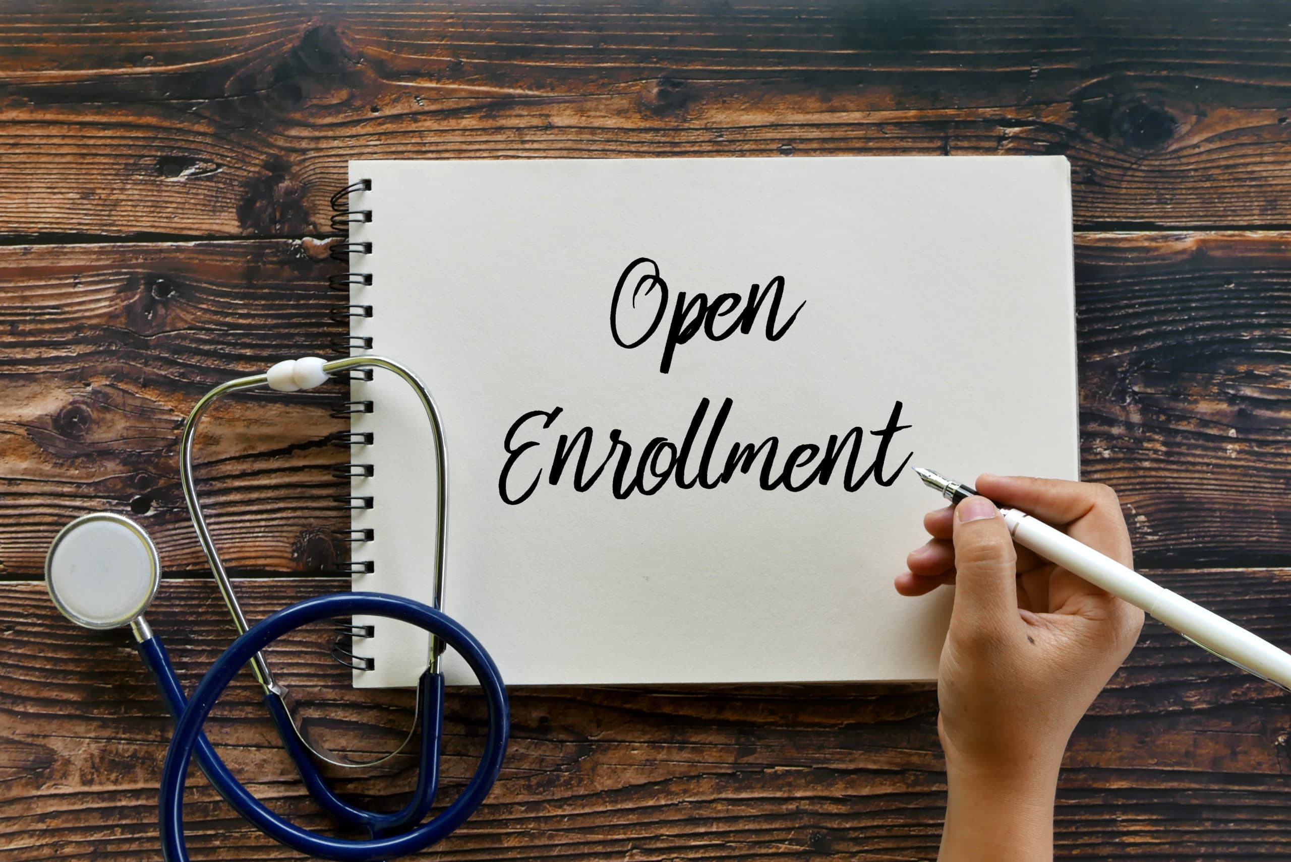 Top view of stethoscope and hand writing Open Enrollment on notebook on wooden background.